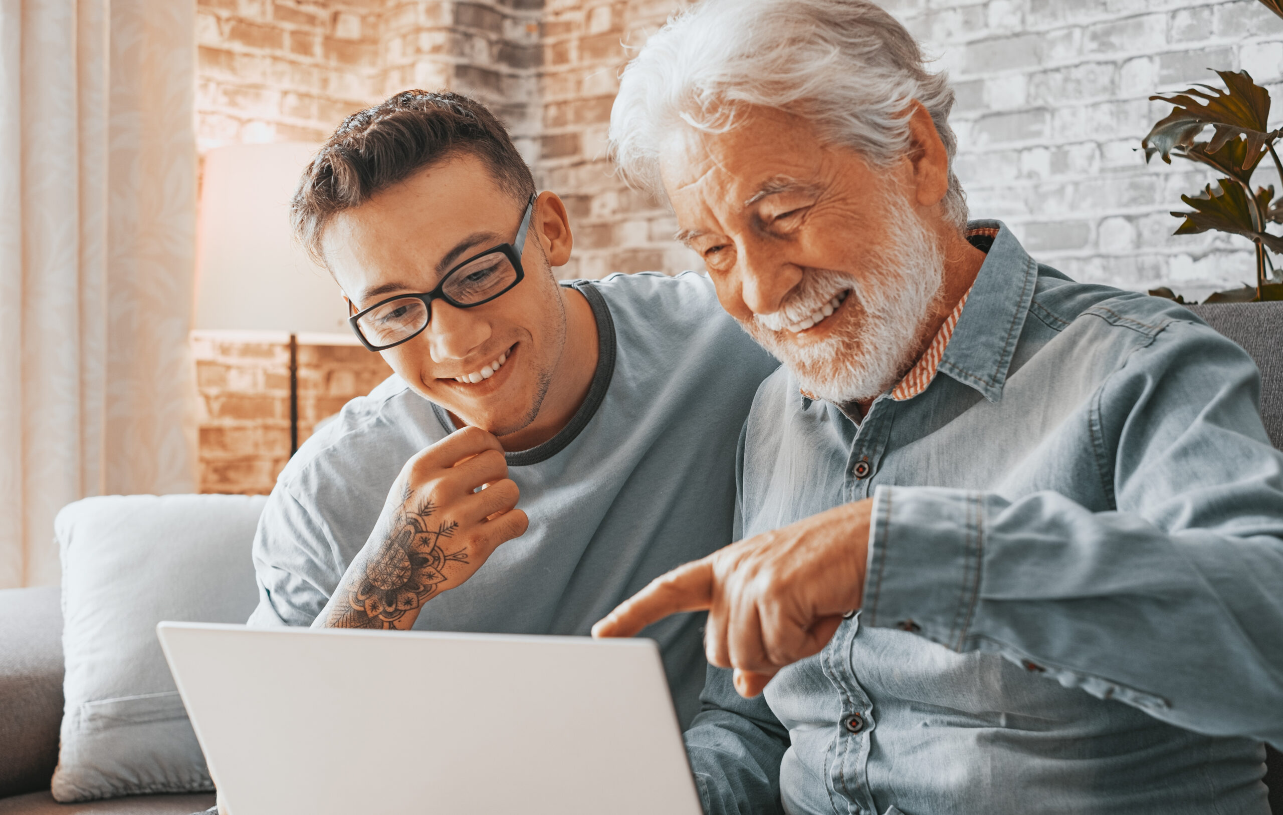 Senior man and young boy with tattoo smiling and looking together at laptop at home. Happy nephew teaching and showing new computer technology to his old grandfather. Old man learn to use computer.