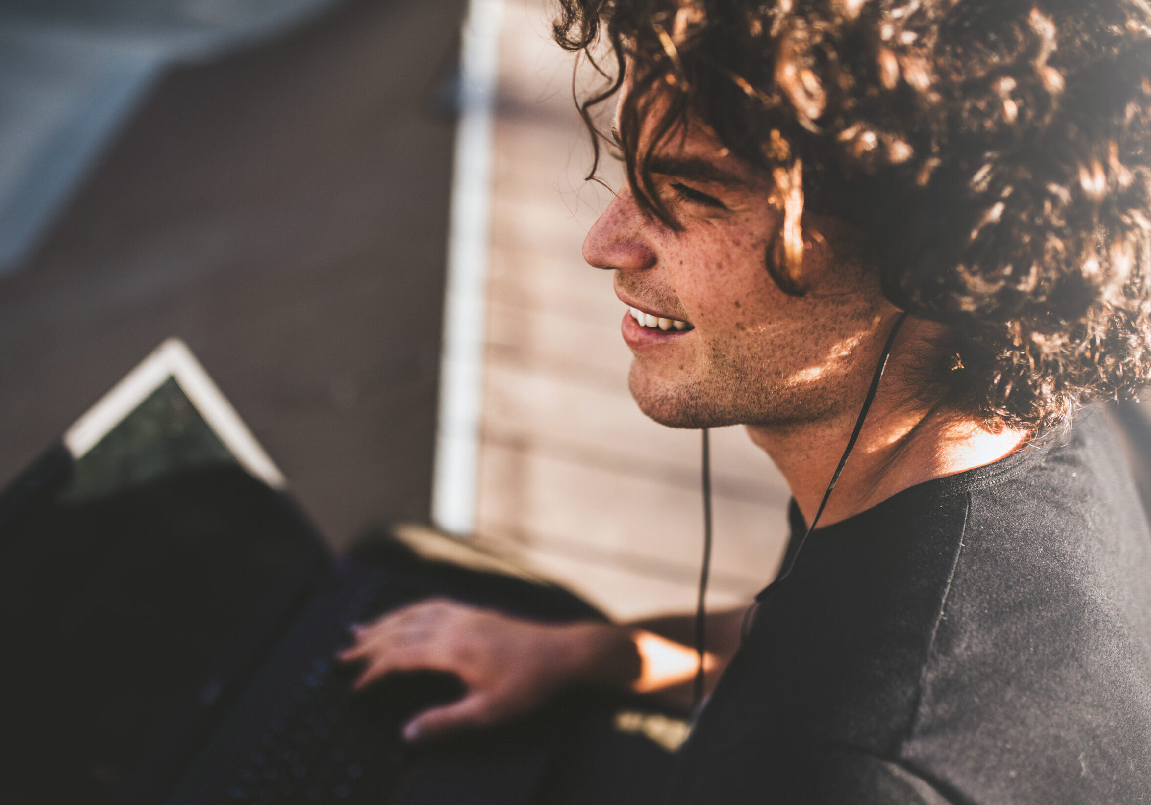Closeup rear view shot of smiling male with curly hair using laptop for chatting online with friends and earphones to listening the music, connected to free wireless. Handsome man texting messages.